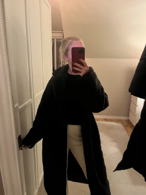 Svart lång teddyjacka med krage - Mysig svart teddyjacka i lång modell med bred krage och avslappnad passform. Jackan är oversized och har långa ärmar, perfekt för kalla dagar. Den fluffiga ytan ger en cool och trendig vibe.