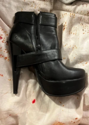 Svarta stövletter med platå och klack - Säljer ett par svarta stövletter med hög klack och rejäl platåsula. Skorna har rund tå, dragkedja på sidan och ett dekorativt spänne upptill. En stilren och edgy look, perfekta för dig som gillar att sticka ut. Dom har små skador men det syns inte när du har på dom!