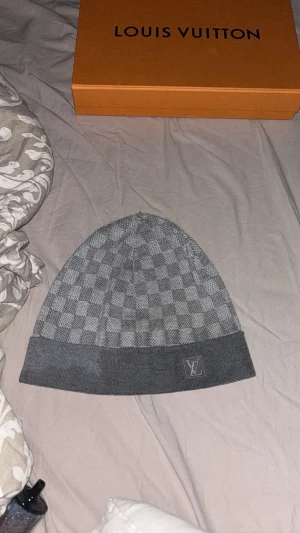 Grå Louis Vuitton rutig mössa - Snygg grå mössa från Louis Vuitton med klassiskt rutigt mönster och LV-logga framtill. Mössan är stickad och har en bred uppvikt kant. Perfekt accessoar för dig som vill ha en lyxig och stilren look under kyliga dagar. Kom med prisförslag 