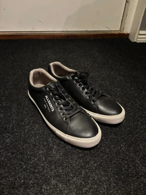 Svarta sneakers från AllSaints - Svarta sneakers från AllSaints med vit sula och klassisk snörning. Skorna har rund tå och är tillverkade i skinn med vit logotyp på sidan. Snygg kontrast mellan svart ovandel och vit yttersula med mönstrad undersida. Perfekt för dig som gillar stilrena och moderna sneakers.