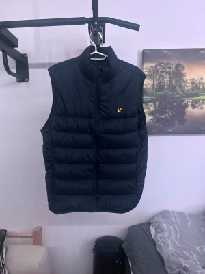 Marinblå dunväst från Lyle & Scott M - Snygg marinblå dunväst från Lyle & Scott i storlek M. Västen har quiltad design, gul logotyp på bröstet och dragkedja framtill. Perfekt för lager-på-lager och har en stilren, sportig look. Materialet är polyester och dun, vilket gör den lätt och varm.