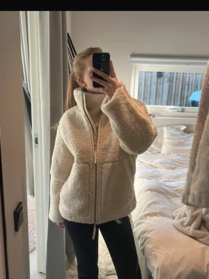 Beige teddyjacka med dragkedja - Supermysig teddyjacka i beige med hög krage och hel dragkedja framtill. Jackan har två stora fickor och en loose passform som ger en chill vibe. Perfekt för kalla dagar när du vill vara både varm och snygg. Materialet är fluffigt fleece och jackan är långärmad.