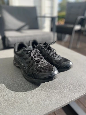 Svarta Asics sneakers  - Säljer ett par svarta Asics sneakers med snyggt mönstrad mesh och detaljer i syntet. Skorna har snörning och en platt sula som ger bra komfort. Perfekt för dig som gillar sportig stil och vill ha ett par schyssta sneakers till vardagen.