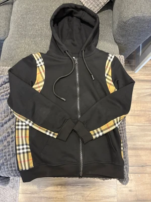 Svart Burberry hoodie med rutig detalj - Svart hoodie från Burberry med klassiska rutiga paneler i beige, svart, vit och röd längs ärmar och axlar. Tröjan har huva med snörning, hel dragkedja framtill och två fickor. Snygg streetstil med exklusiv känsla och tydlig logga på snörningen.