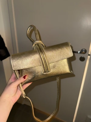 Latalia guldig väska  - Säljer denna fina guldiga väska ifrån märket Latalia i 100% läder. Unik väska som kan bäras både som crossbody eller clutch, man kan även hålla den i ”snöret” så blir det nästan som armband också,också med tanke på att väskan är guldig.Det finns ett fack med dragkedja och ett annat fack i väskan. Perfekt till balen, som julklapp eller nyår.Endast testad.Nypris är 990 kr. Se bilder,skötselråd och mer info på bilderna och/eller på deras egna hemsida.Kolla gärna in mina andra annonser, samfraktar!🥰
