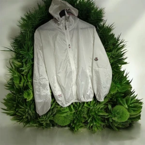 Vit tunn jacka med huva - Beskrivning: Lätt och tunn vit jacka i glansigt material. Har huva med justerbara snören, hel dragkedja framtill och elastiska muddar. En liten tygpatch sitter ej äkta  på ena ärmen. Jackan är helt ny med tag kvar. 