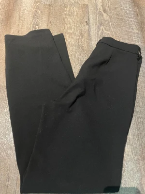 Svarta kostymbyxor från Vero Moda - Snygga svarta kostymbyxor från Vero Moda med klassisk rak passform. Byxorna har dragkedja och knappstängning i midjan, vilket ger en stilren look. Perfekta för dig som gillar enkel och clean stil.