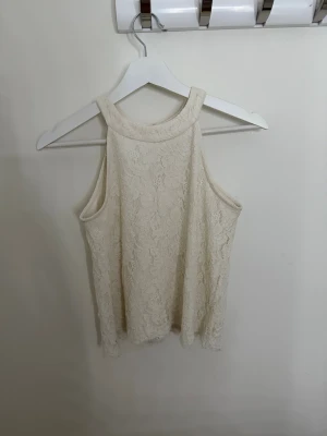 Spetsblus i ljus beige från Lindex - Säljer en superfin ljus beige blus från Lindex i storlek 146/152. Blusen är ärmlös och har ett blommigt spetsmönster över hela plagget. Rund halsringning och två pärlknappar bak i nacken för extra detalj. 