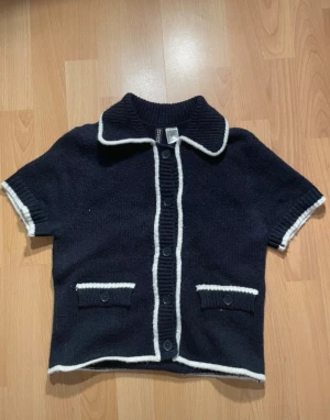 Mörkblå stickad kofta från H&M - Mörkblå stickad kofta från H&M med vit kant längs krage, ärmslut, fickor och knappslå. Kortärmad modell med klassisk krage och dekorativa knappar framtill. 