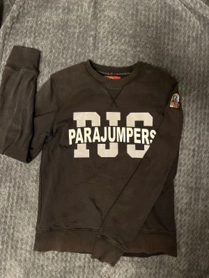 Parajumpers kofta  - Svart långärmad fleece-tröja från Parajumpers med stor vit print 'PJS PARAJUMPERS' på bröstet och patch på ärmen. Rund hals, ribbade muddar och nederkant. Perfekt för dig som gillar streetwear och vill ha en skön och snygg tröja. 
