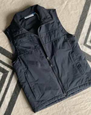 Tunn väst från Race Marine - Svart väst från Race Marine med krage och dragkedja framtill. Fickor på båda sidor med dragkedja och diskret broderad logga på bröstet. Snygg och enkel design i syntetmaterial, perfekt att slänga över en hoodie eller tröja.