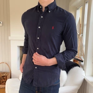 Mörkblå skjorta från Ralph Lauren small slim fit - Snygg mörkblå skjorta från Ralph Lauren i slim fit-modell. Skjortan har klassisk krage med knappar, vit knappstängning framtill och ett rött broderat logomärke på bröstet. Tillverkad i mjuk bomull och har långa ärmar med manschettknappar.