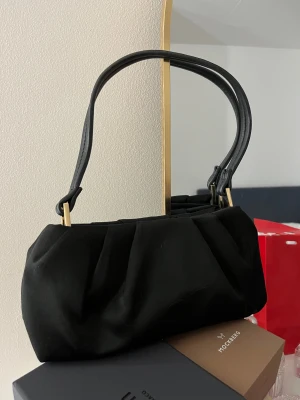 Svart axelväska med gulddetaljer - Snygg svart axelväska med mjuk, veckad design och guldfärgade detaljer vid handtaget. Väskan har ett klassiskt format och stängs med dragkedja. Perfekt storlek för det viktigaste och passar till många olika stilar. Pris går att diskuteras