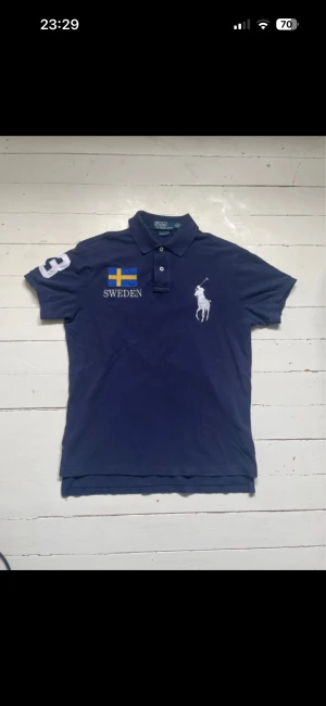 Marinblå pikétröja Polo Ralph Lauren - Snygg marinblå pikétröja från Polo Ralph Lauren med broderad svensk flagga och texten SWEDEN på både fram- och baksidan. Stor vit siffra 3 på ärmen och ryggen, samt klassisk polospelare-logga. Tröjan är i bomull och har en normal passform med korta ärmar.
