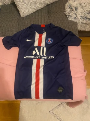 PSG matchtröja Nike L - Paris Saint-Germain fotbollströja i marinblått med vita och röda vertikala ränder på framsidan. Tröjan har korta ärmar, rund hals och PSG-logga på bröstet. Materialet är lätt och andas, perfekt för träning eller match. Snygg Nike Swoosh och sponsortryck på framsidan.