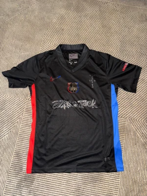 Svart FC Barcelona x Nike fotbollströja - Säljer en svart FC Barcelona fotbollströja från Nike med klubbmärke på bröstet och röda samt blå detaljer på sidorna. Tröjan har korta ärmar, V-ringad krage och tryck på bröstet. Materialet är lätt och andas, perfekt för match eller träning. Det finns en i storlek M och en i storlek XXL men passar bra för XL eller L
