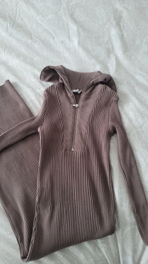 Ribbad långklänning med dragkedja från Zara - Säljer en ribbad långklänning från Zara i en snygg brun nyans. Klänningen har lång ärm, huva och en cool dragkedja framtill. Materialet är mjukt och stretchigt, perfekt för dig som gillar en slim passform och stilren look.