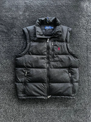 Ralph Lauren väst - Svart dunväst från Polo Ralph Lauren med röd broderad logga på bröstet. Västen har hög krage, tre praktiska fickor med dragkedja och quiltad design. Perfekt för lager-på-lager och har en clean, sportig look. Skick 7/10, hål innuti fickan men inget man ser på utsidan, storlek S men passar mer som M. Pris går att diskutera✅