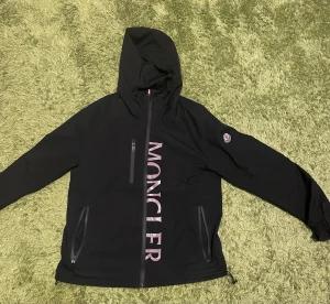 Svart Moncler vindjacka med huva - Svart vindjacka från Moncler med stor logotyp i rosa på framsidan och klassisk Moncler-patch på ärmen. Jackan har huva, tre dragkedjefickor och justerbara muddar. Materialet är syntet och dun, perfekt för kyliga dagar. Snygg och sportig look med clean design.