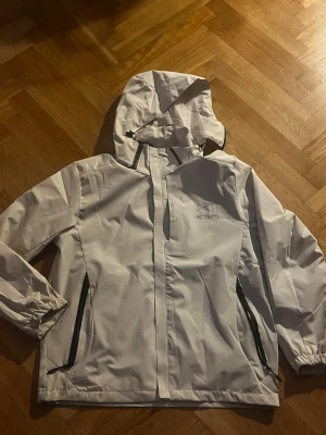 Vit Arc'teryx regnjacka med huva - Snygg vit regnjacka från Arc'teryx med huva och svarta dragkedjor. Jackan har stilren design, justerbara ärmslut och två praktiska fickor med dragkedja. Perfekt för blöta dagar och outdoor-äventyr. Logga på bröstet och detaljer på ryggen.