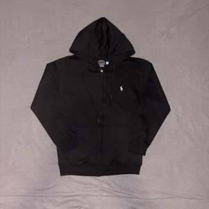Svart hoodie från Polo Ralph Lauren - Svart hoodie från Polo Ralph Lauren i slim fit med klassisk huva och snörning. Tröjan har dragkedja framtill, ribbade muddar och fickor. Ikoniska Polo-loggan broderad i vitt på bröstet. Perfekt för chill dagar och streetwear.
