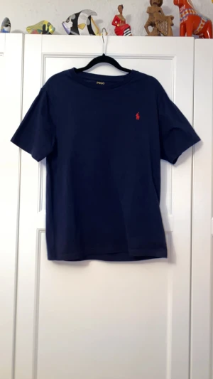 Mörkblå t-shirt från Polo Ralph Lauren - Snygg mörkblå t-shirt från Polo Ralph Lauren med röd logga på bröstet. Modellen är kortärmad och har en rund hals. Tillverkad i mjuk bomull som känns skön mot huden. Perfekt för dig som gillar stilrena och tidlösa plagg. Den är lite stor i storleken så den passar även S och i vissa fall M i män storlek.