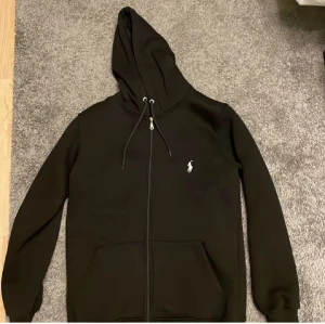 Svart hoodie från Polo Ralph Lauren - Svart hoodie från Polo Ralph Lauren med dragkedja, snörning vid huvan och klassisk logga broderad i vitt på bröstet. Tröjan har långa ärmar och två fickor framtill. Perfekt för chill dagar och streetstyle.