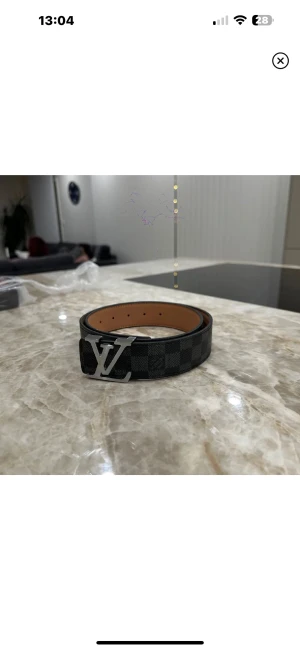 Bälte - Säljer ett bälte i svart och grått med klassiskt rutigt mönster och en stor silverfärgad -logga som spänne. Bältet är tillverkat i skinn och har en stilren, bred design som ger en lyxig vibe. Perfekt för dig som gillar exklusiva accessoarer.