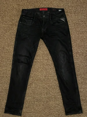 Replay Anbass svarta  jeans - Svarta Replay Anbass jeans med klassisk femficksdesign. Jeansen har en mörk tvätt och subtila slitningar, med Replay-logga på fickan och bakre läderpatch. Perfekt för dig som gillar en smal siluett och stilren look.