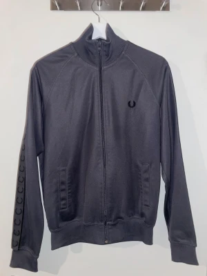 Fred Perry zip - Säljer en mörkgrå track jacket från Fred Perry med svart logga på bröstet och ikoniska laurel wreath-band längs ärmarna. Jackan har hög krage, hel dragkedja och ribbade muddar. Tillverkad i mjuk polyester för en sportig och clean look.