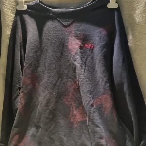 Svart långärmad tröja från BOSS - Svart långärmad tröja från BOSS med diskret röd logga på bröstet och grönt märke i nacken. Tröjan har mörkröda detaljer och klassisk rund halsringning. Perfekt för dig som gillar stilrena och bekväma plagg med en twist.