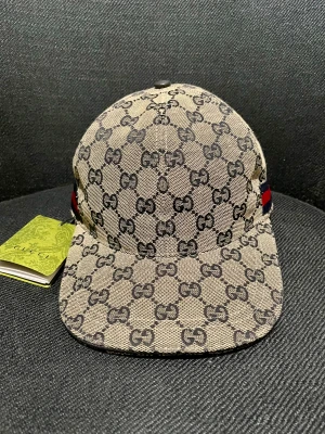 Gucci keps  - Säljer en snygg keps från Gucci med klassiskt GG-monogram i blått på grå botten. Kepsen har böjd skärm och ikoniska röda och blå ränder på sidan. Tillverkad i bomullscanvas med mörkblått innerfoder och märkets logga invändigt.