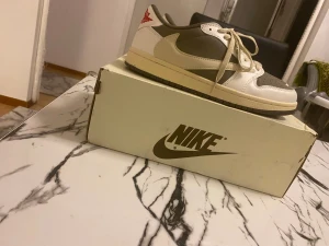 Nike Air Jordan 1 Low Travis Scott - Nike Air Jordan 1 Low Travis Scott i beige och mörkgrått med röda detaljer på hälen. Snygg kontrast mellan vitt skinn och mörkgrå mocka, klassisk snörning och rund tå. Ikoniska loggor på tungan och hälen, samt Travis Scott smiley. Perfekt för dig som vill sticka ut med hypade sneakers. Köpt för 2900kr.