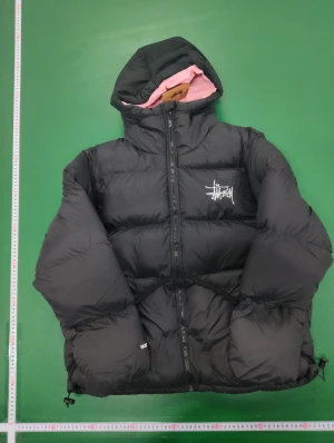 Svart pufferjacka från Stüssy M - Svart pufferjacka från Stüssy med rosa foder och huva. Jackan har Stüssy-logga på bröstet, dragkedja framtill och vattenavvisande yta. Den är quiltad och har en loose passform, perfekt för kalla dagar. Snygg streetwear-vibe och riktigt skön att bära.