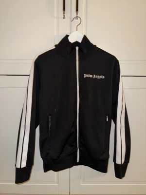 Palm Angels svart zip-tröja - Svart zip-tröja från Palm Angels med vita ränder längs ärmarna och logga på bröstet. Hög krage, hel dragkedja och två fickor med dragkedja. Sportig och clean look som funkar till streetwear. Materialet känns mjukt och har lite stretch.