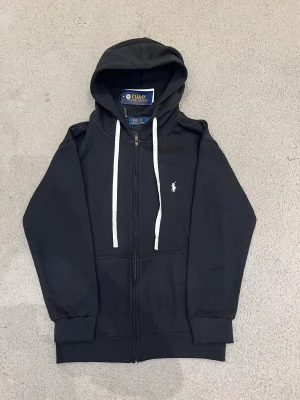 Ralph Lauren Kofta  - Svart hoodie från Polo Ralph Lauren med vit broderad logga på bröstet, vita snören i huvan och dragkedja framtill. Tröjan har ribbade muddar och två fickor fram. Perfekt för chill dagar och enkel att matcha med jeans eller joggers.