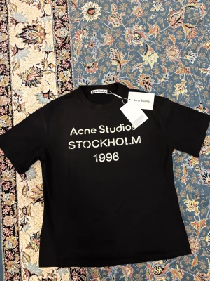 Svart Acne Studios t-shirt Stockholm - Svart t-shirt från Acne Studios med vit text 'Acne Studios STOCKHOLM 1996' Helt ny  Klassisk rund hals och korta ärmar. Tillverkad i mjuk bomull för en skön och avslappnad känsla. Perfekt för dig som gillar stilrena och trendiga plagg.
