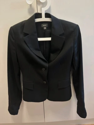 Svart randig kavaj från Mexx - Snygg svart kavaj från Mexx med diskreta tunna ränder. Klassisk modell med två knappar framtill, spetsiga slag och två fickor med lock. Perfekt för dig som gillar stilrena och tidlösa plagg