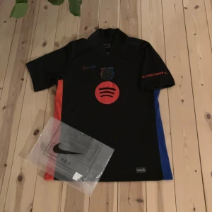 Svart FC Barcelona Nike fotbollströja - FC Barcelona fotbollströja från Nike i svart med röda och blå detaljer på sidorna. Tröjan har korta ärmar, Dri-FIT-material och tryck med Spotify-logga, klubbmärke och sponsortryck på ärmen. Perfekt för dig som vill sticka ut på planen eller läktaren.