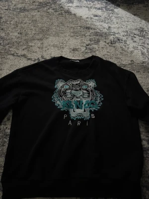 Svart Kenzo sweatshirt med tiger - Svart sweatshirt från Kenzo med ikonisk broderad tiger och texten 'Kenzo Paris' i turkos och grått framtill. Tröjan är i 100% bomull och har rund hals samt långa ärmar. Perfekt för dig som gillar streetwear och vill sticka ut med ett snyggt statement-plagg.
