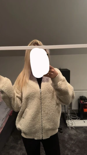 Beige teddyjacka med dragkedja - Mysig beige teddyjacka med fluffig yta och ribbade muddar. Jackan har dragkedja framtill och två fickor på sidorna. Perfekt för kyliga dagar och har en loose passform som ger en avslappnad vibe.
