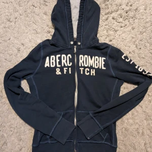 Abercrombie & Fitch Zip hoodie - Snygg marinblå hoodie från Abercrombie & Fitch med vit broderad logga på bröstet och ärmen. Tröjan har huva med dragsko, hel dragkedja framtill och två fickor. Stl M men passar perfekt till stl S också. Längd: 56 cm Bredd: 45 cm