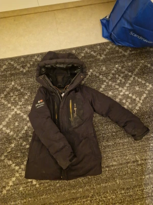 Svart damjacka från Sail Racing - Svart dunjacka från Sail Racing med huva och flera praktiska fickor med gula dragkedjor. Jackan har lång ärm, quiltad insida och snygga logodetaljer på bröstet och ärmen. Perfekt för kalla dagar och har en clean, sportig look.