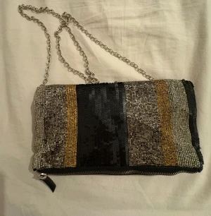 Randig paljettväska med kedja - Snygg axelväska med silverfärgad kedja och dragkedja. Framsidan har breda ränder i svart, guld och silver med paljetter, medan baksidan är i svart skinn. Perfekt storlek för det viktigaste och ger en glittrig touch till din outfit.