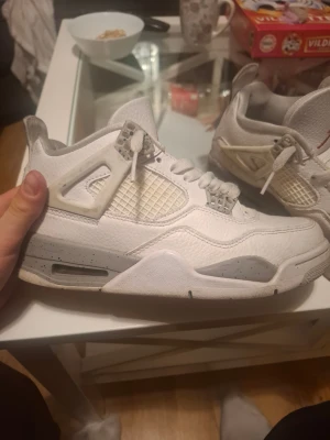 Nike Air Jordan 4 White Cement sneakers - Säljer ett par Nike Air Jordan 4 White Cement sneakers i vitt skinn med grå och svarta detaljer. Klassisk meshpanel på sidorna, grå stänkmönstrad sula och ikonisk röd Jumpman-logga på plösen. Skorna har snörning och en chunky siluett som sticker ut.
