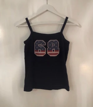 Marinblå linne med tryck från Two 2U You - Snygg marinblå linne från Two 2U You med smala axelband och stort tryck med siffran 68 framtill i amerikanskt flaggmönster. En tråd har lossnat i kanten, framgår på bild