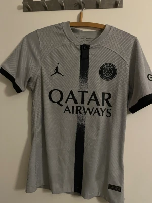 PSG x Jordan grå fotbollströja - Snygg PSG x Jordan fotbollströja i ljusgrått med svart tryck och detaljer. Kortärmad med svart kant vid ärmslut, diskret geometriskt mönster i tyget och klassisk rund hals. Materialet är lätt och andas, perfekt för träning eller match. Qatar Airways-logga på bröstet.