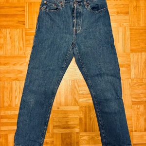 Levi's 501 blå jeans W28 L28 - Klassiska Levi's 501 jeans i blå denim med raka ben och knappgylf. Modellen har fem fickor, hög midja och snygga kontrastsömmar. Perfekt för dig som gillar tidlös stil och vill ha ett par jeans som alltid funkar. Märke och storlek syns tydligt på läderpatchen bak.