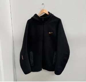 Svart Nike NOCTA hoodie med huva - Svart hoodie från Nike NOCTA med gul Swoosh på bröstet och NOCTA-tryck på ärmen. Tröjan har huva, dragkedja framtill och två fickor med zip. Materialet är mjukt och känns som bomull och polyester. Perfekt för dig som gillar streetwear och snygga detaljer.