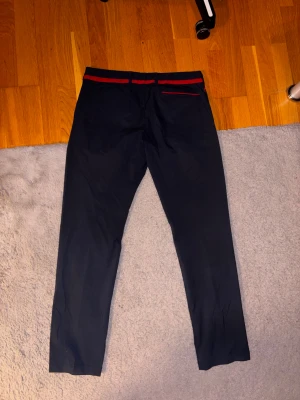 Mörkblå kostymbyxor Hugo Boss - Snygga mörkblå kostymbyxor från Hugo Boss med extra slim fit-passform. Byxorna har röda detaljer vid midjan och fickorna samt diskret Boss-logga på sidan. Materialet är mjukt och har en lyxig känsla, perfekt för dig som gillar stilrena och moderna plagg.